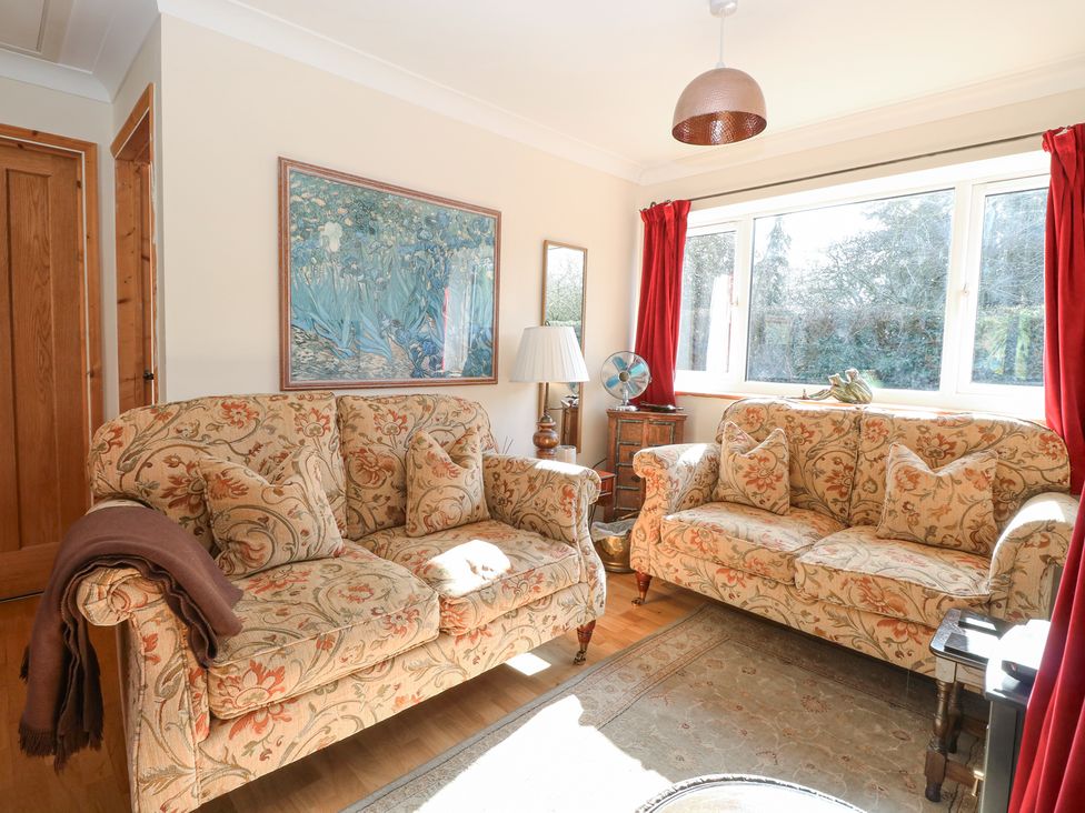 A sitting room with two patterned sofas a table lamp and a large window with red curtains at The Lodge in Coltishall