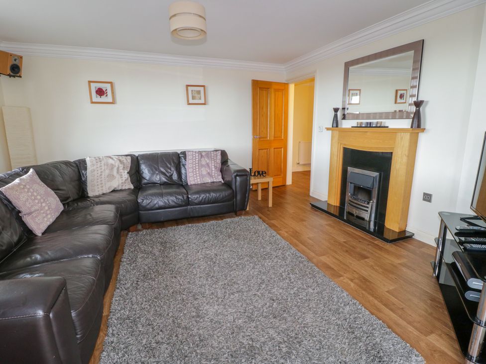 A living room with a black leather corner sofa with cushions a gray rug wooden floor a wooden fireplace with a mirror above and a television on a stand at Lle cyn Braf in Amlwch