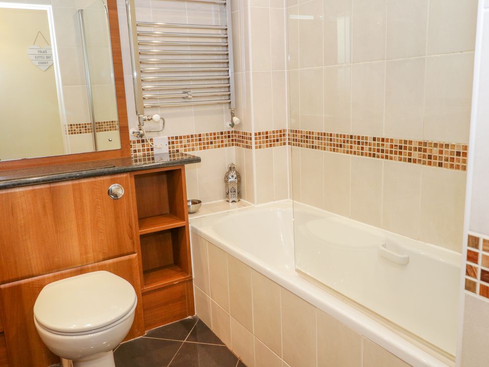A bathroom with a toilet wooden cabinet a bathtub with glass screen and tiled walls at Llecyn Braf in Amlwch