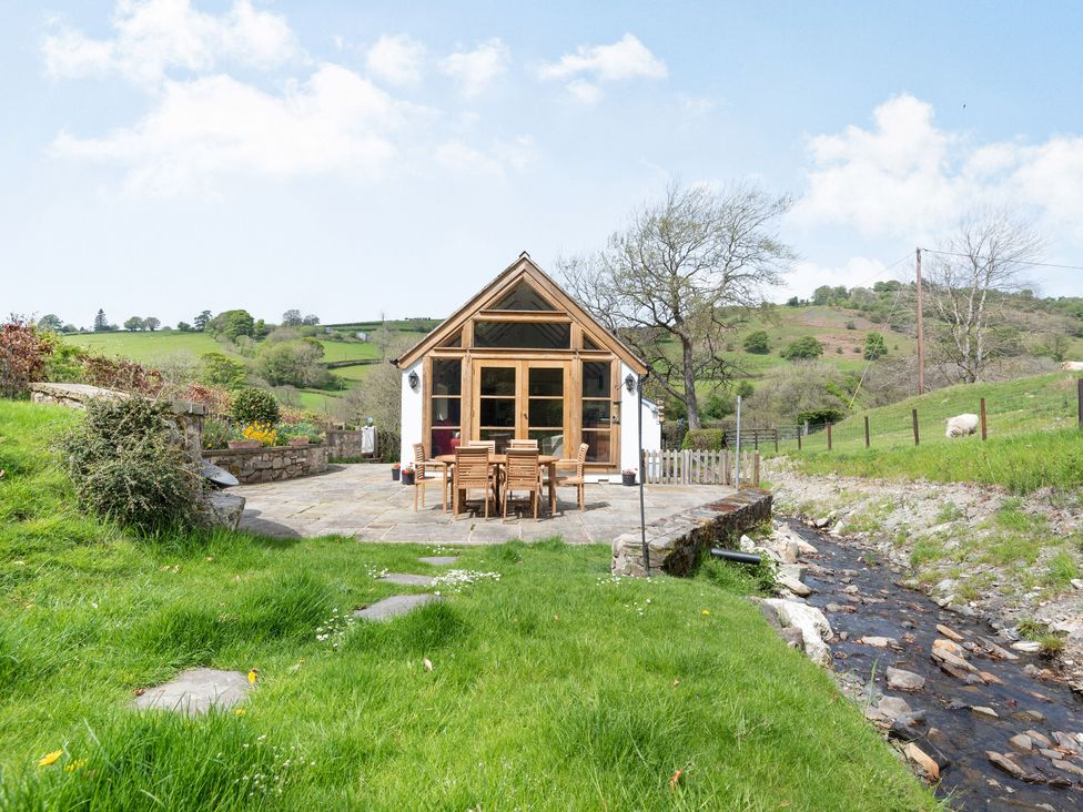 A small house with a wooden patio set outside next to a stream and green hills at Pen Y Bont in Glyn Ceiriog