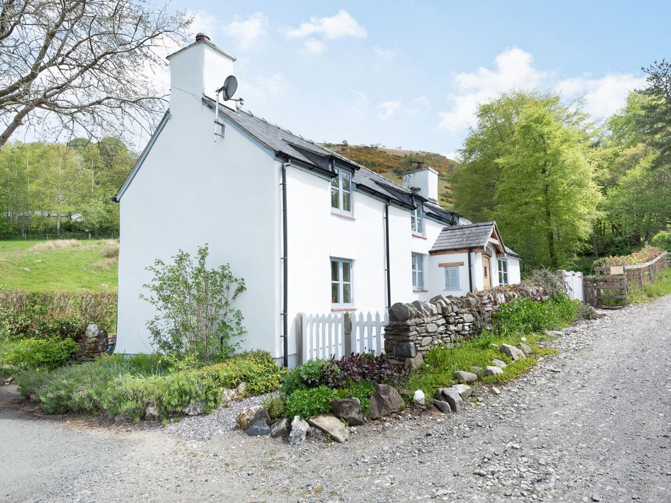 A white two-story cottage with stone wall and garden along a gravel driveway at Pen Y Bont in Glyn Ceiriog