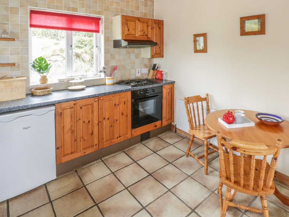 A kitchen with wooden cabinets a black countertop a small white fridge a stove a round wooden table with two chairs and framed pictures on the wall at Seaspray in Tully County Galway