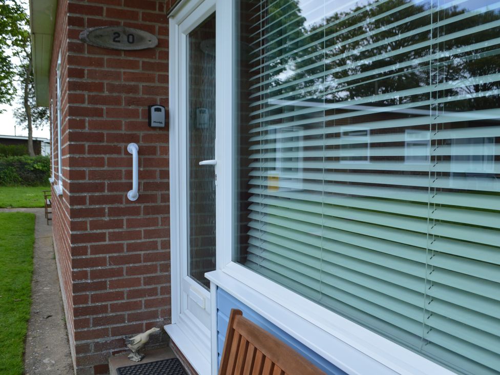 An exterior view of a brick house with a white door and window with blinds showing a wooden bench and a small decorative duck at 20 Siesta Mar Mundesley