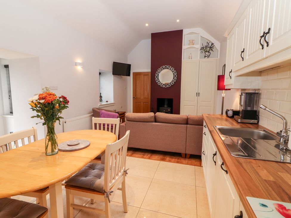 A kitchen and dining area with a wooden table and chairs a vase with flowers on the table a sofa TV and cupboards at Laharn Cottage in Killorglin County Kerry