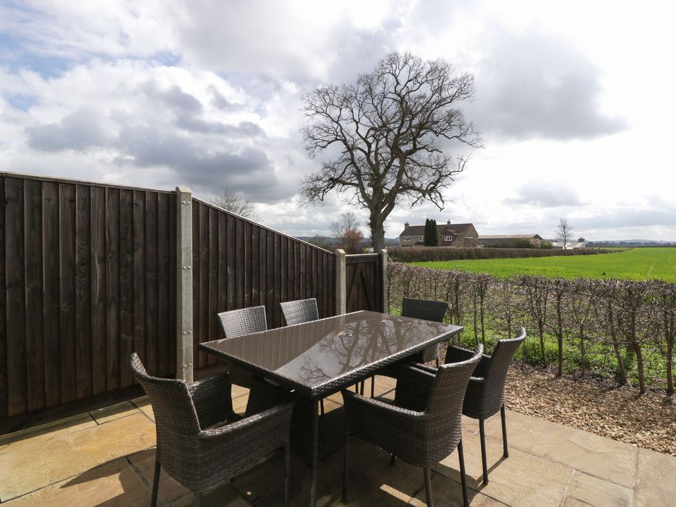 An outdoor patio with a glass table and six wicker chairs near a wooden fence and a hedge with a large tree and fields beyond at Rectory Farm View in Queen Camel near Yeovil