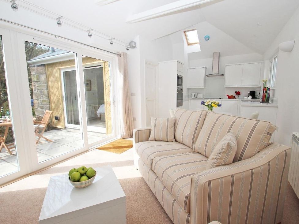 A living area with a striped sofa and bowl of green apples on a table next to a kitchen with white cabinets at Cot Valley Cottage in St Just