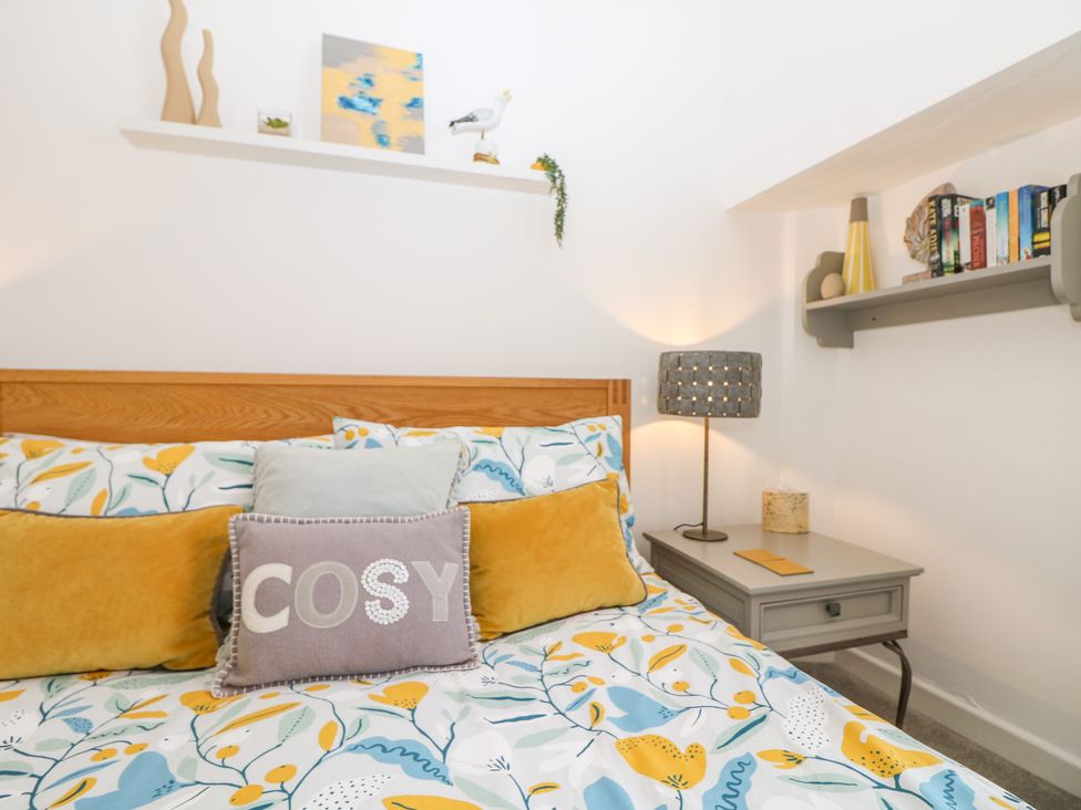 A bedroom with a wooden bed frame colorful bedding cushions and a nightstand with a lamp and books at Sea Breeze in Marazion
