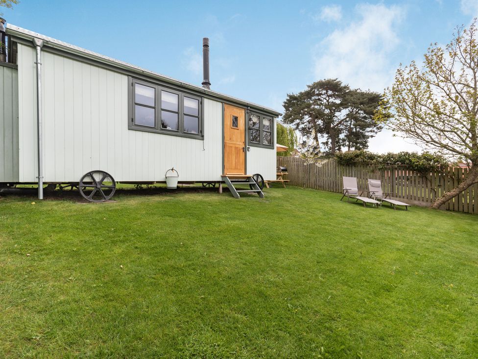 A small cabin on wheels with a wooden door and black windows on a grassy lawn with two lounge chairs near a wooden fence at The Brae in Snainton