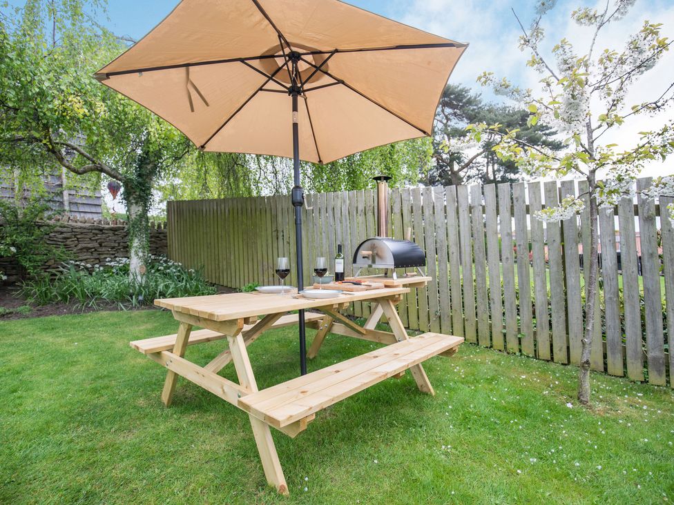 A wooden picnic table with benches and an umbrella with wine glasses and a small outdoor oven in a garden at The Brae in Snainton