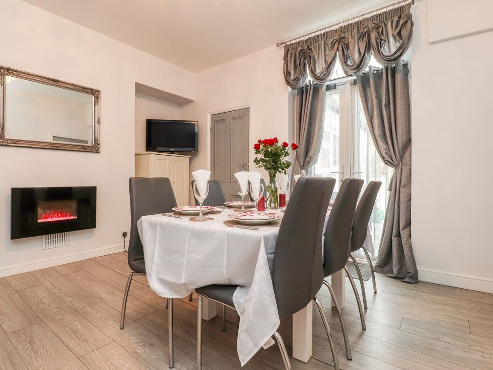 A dining room with a table set for six, a wall-mounted electric fireplace, a mirror, a television on a cabinet, and large windows with draped curtains at Angel Cottage in Skipton
