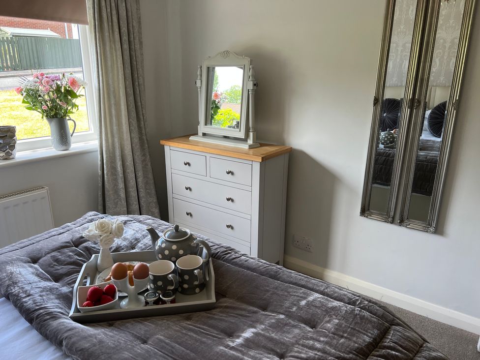 A bedroom with a bed, grey comforter, a tray with tea set and breakfast items, a chest of drawers with a mirror, a window with flowers on the sill and two vertical mirrors on the wall at Araucaria House in Whitby