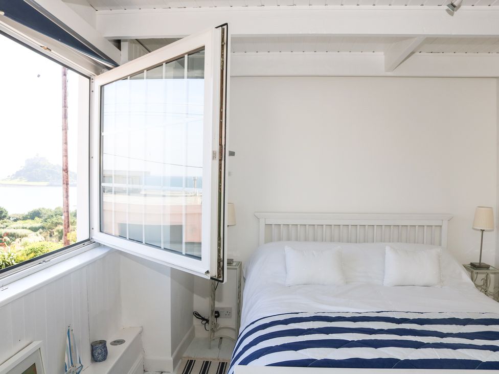 A bedroom with an open window showing a view outside and a bed with pillows and a striped blanket at Ner Sea in Marazion