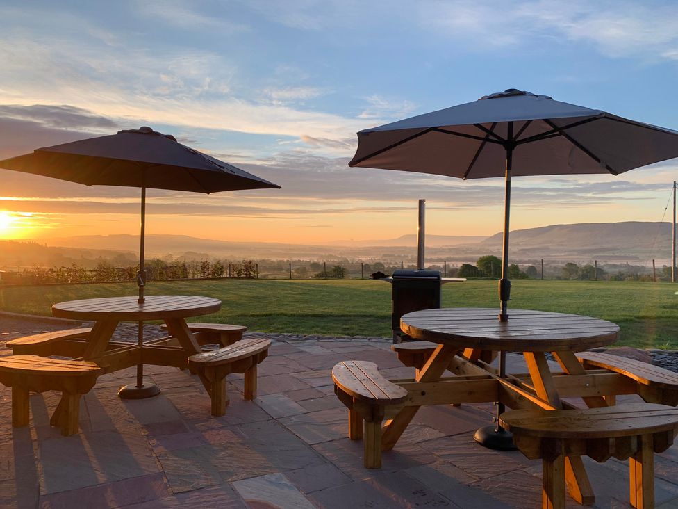 Two wooden picnic tables with umbrellas on a stone patio overlooking a grassy field and hills at sunrise at Cuthbert Hill Farm in Chipping