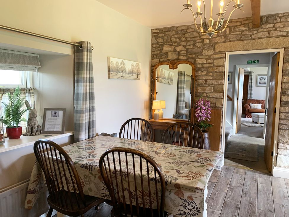 A dining room with a table covered by a patterned tablecloth and four chairs a window with plaid curtains a wooden floor a stone wall a wooden mirror with a lamp and flowers a view into a room with an armchair at Cuthbert Hill Farm in Chipping