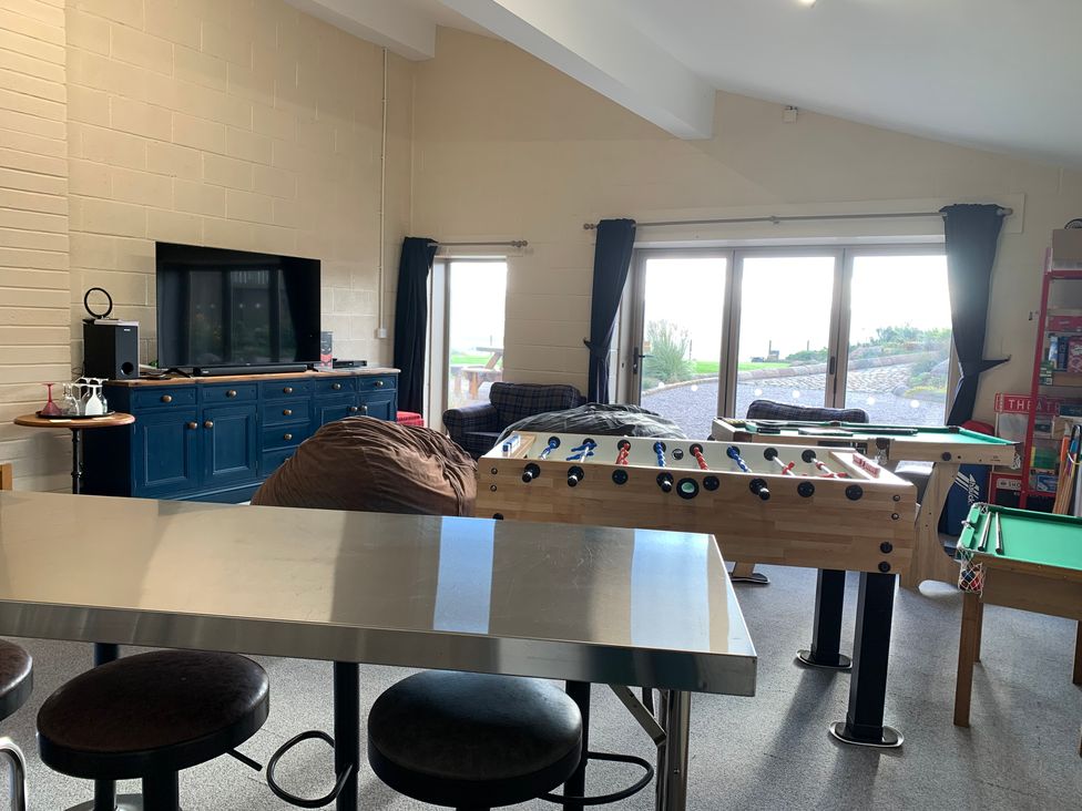 A game room with a foosball table, pool tables, tv on a blue cabinet, sofas, stools, and large windows at Cuthbert Hill Farm in Chipping