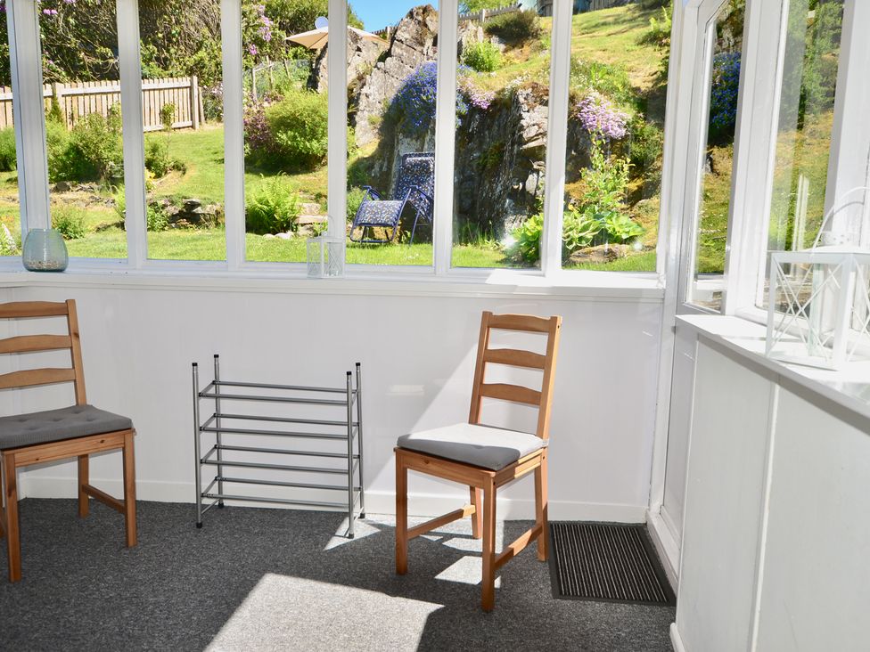 A sunroom with wooden chairs and a small metal rack overlooking a garden at Ling Fell Cottage in Backbarrow