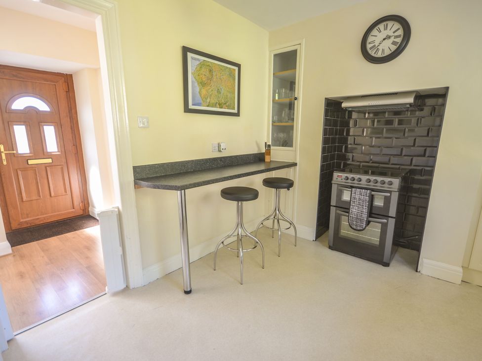 A kitchen with a stove a black tiled backsplash a wall clock a black countertop with two stools and a wooden door at Ling Fell Cottage in Backbarrow