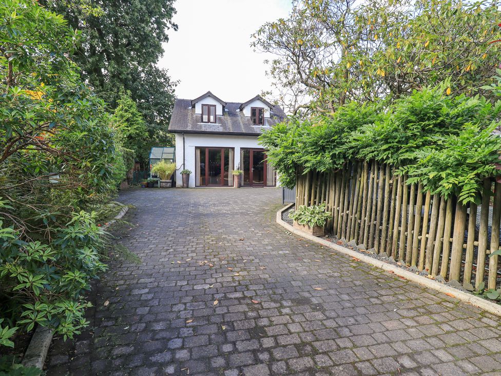 A paved driveway leading to a two-story house with dormer windows and large glass doors bordered by greenery and wooden fencing at Stonegarth Mews in Grasmere