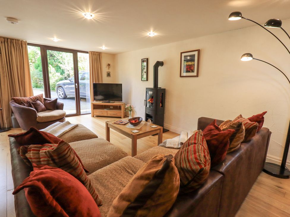A living room with a brown sofa, armchair, wooden coffee table, TV on stand, wood stove, and floor lamp at Stonegarth Mews in Grasmere