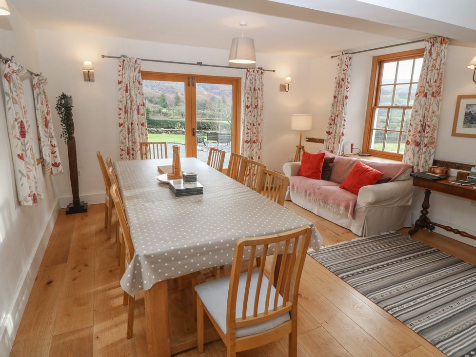 A dining room with a long table covered with a polka dot tablecloth surrounded by wooden chairs a sofa with red pillows a striped rug and curtains covering windows at Peniarth Ganol in Tywyn
