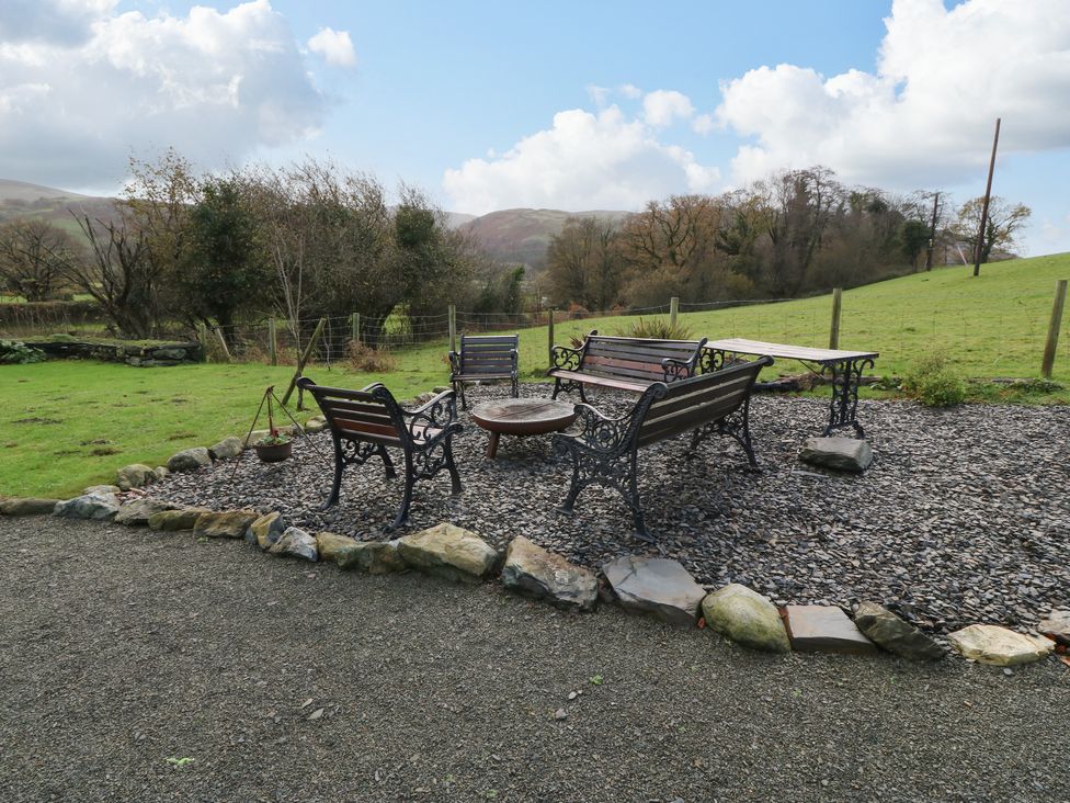 An outdoor seating area with benches and chairs around a fire pit on a gravel surface surrounded by grass and trees at Peniarth Ganol in Tywyn