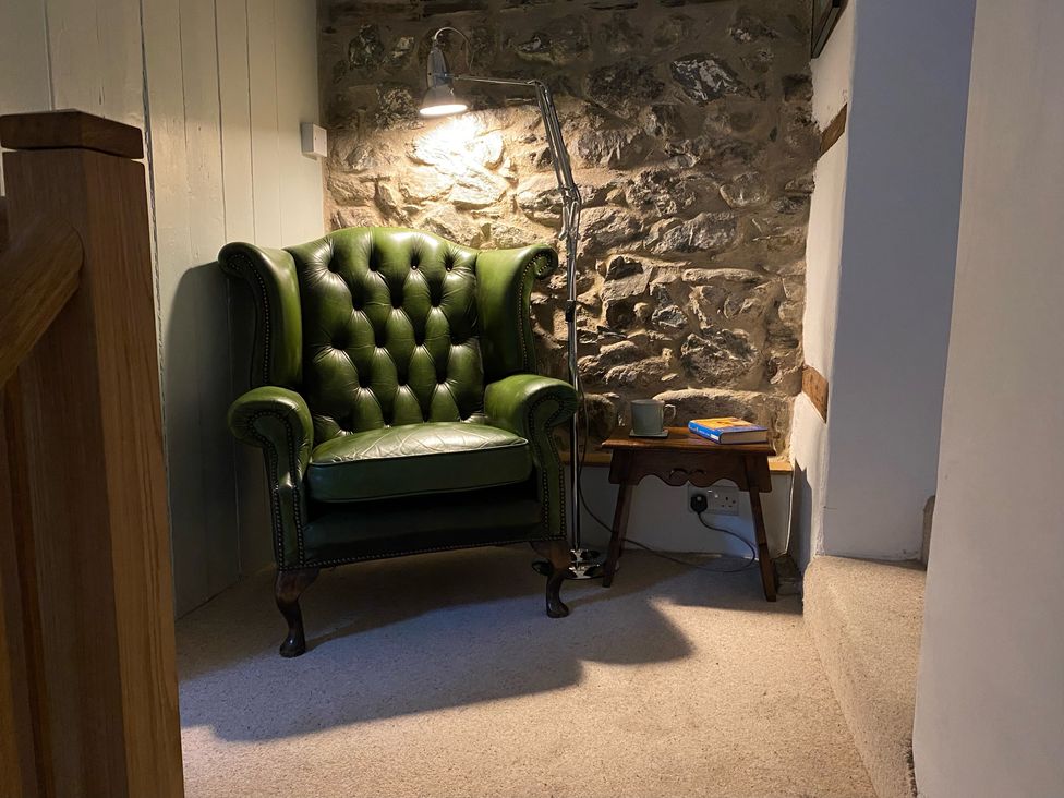 A corner with a green leather armchair a standing lamp and a small wooden side table with a book and mug against a stone wall at Peniarth Ganol in Tywyn