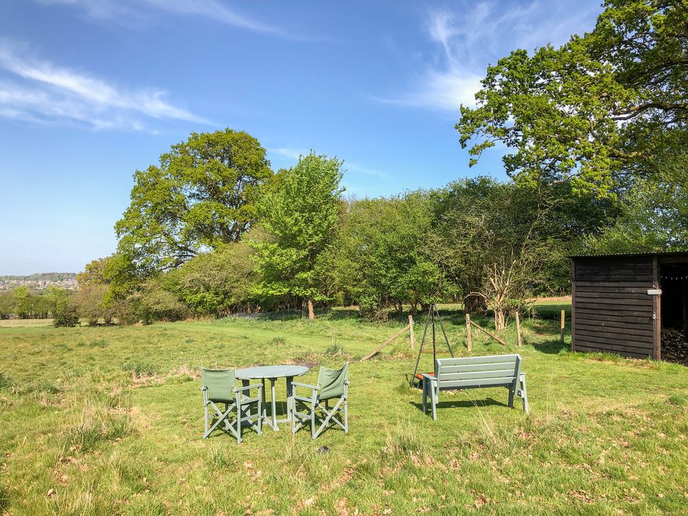 An outdoor grassy area with two green chairs and a table a green bench a wooden shed and trees at Pheasant Lodge in Wimborne Minster