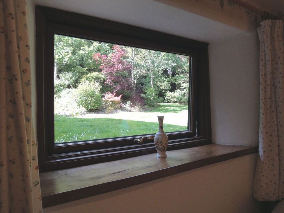 A window with floral curtains and a vase on the wooden windowsill overlooking a garden with trees and grass at Water Barn in Manaton near Lustleigh