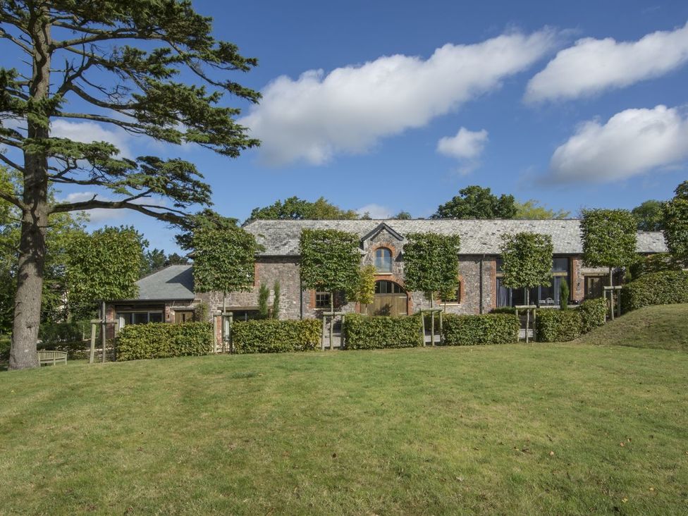 A stone building with a slate roof behind a row of pruned trees and hedges with a grassy lawn and a large tree on the left at Grooms Cottage Talaton near Ottery St Mary