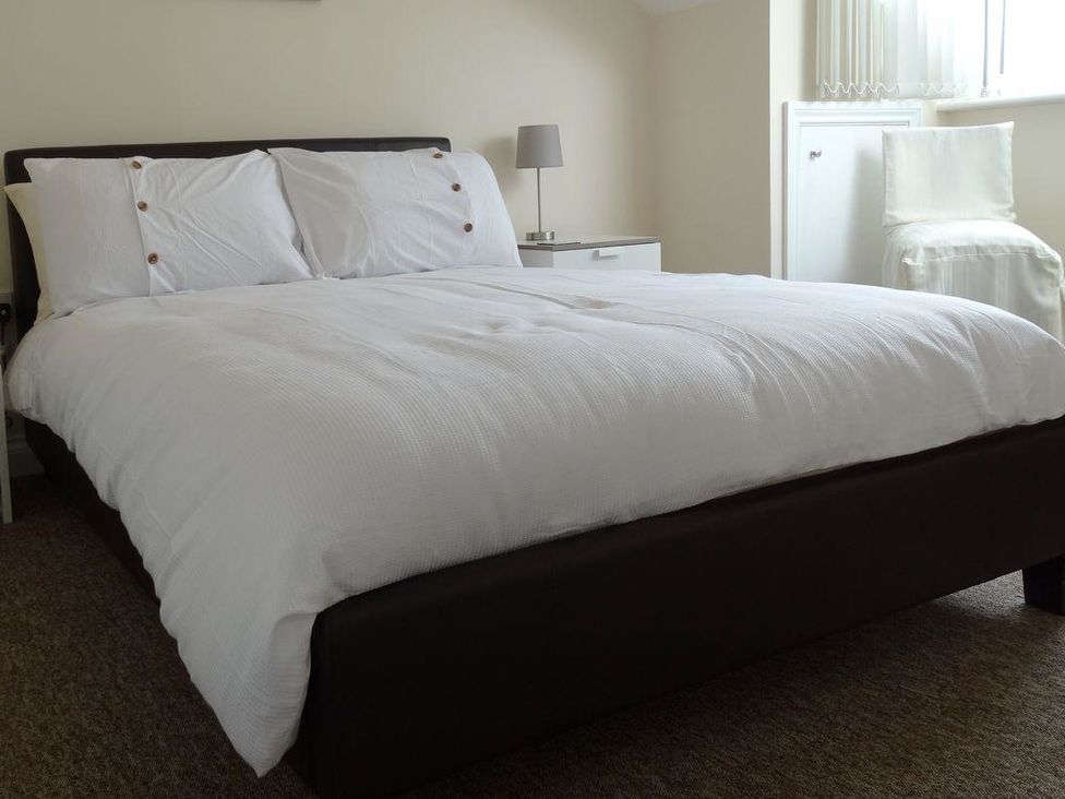 A bedroom with a bed white bedding a bedside table with a lamp and a chair at 2 Carlton Mews in Exmouth