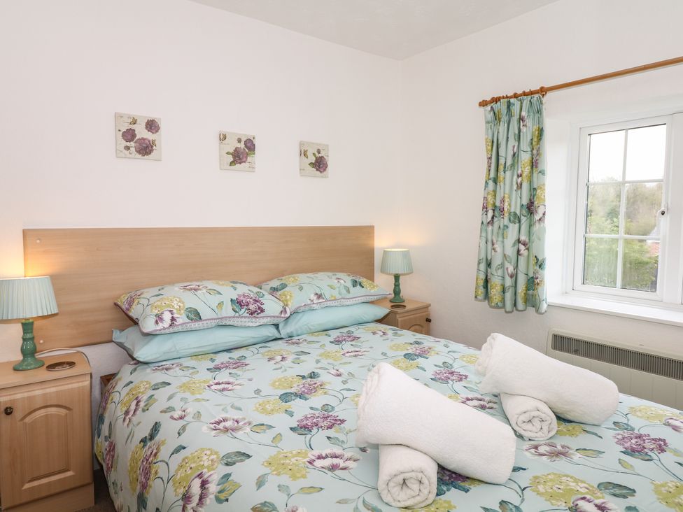 A bedroom with a double bed with floral bedding two bedside tables with lamps a window with floral curtains and rolled towels on the bed at Poppy Cottage in Charlton Marshall