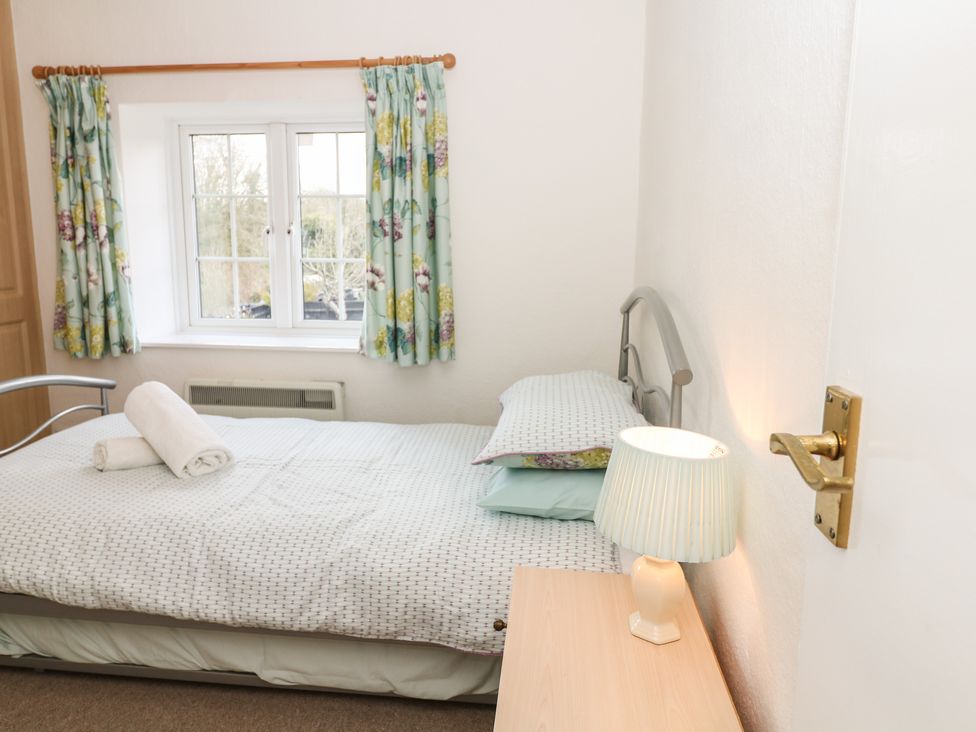 A bedroom with a bed rolled towels bedside table with lamp window with floral curtains at Poppy Cottage in Charlton Marshall