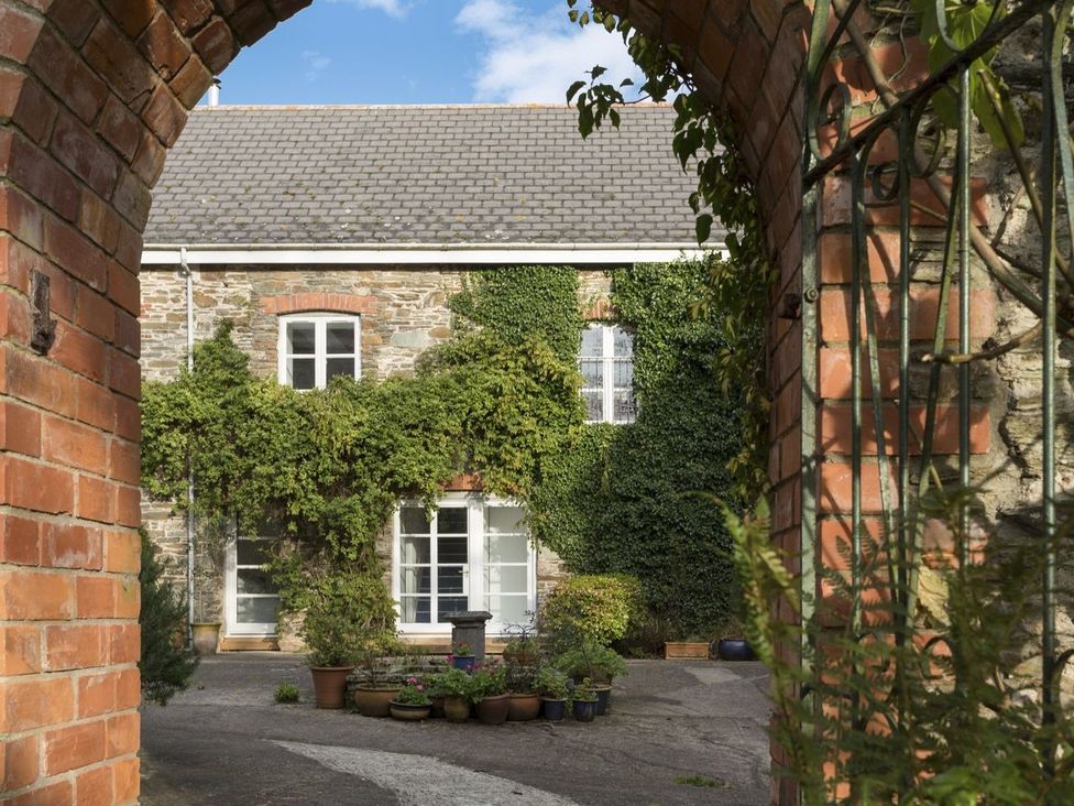 A courtyard with potted plants and a stone house covered with ivy seen through a brick archway at The Gallery Aveton Gifford
