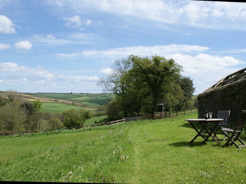 A grassy outdoor area with a wooden table and two chairs near a stone wall with hills and trees in the background at The Gallery in Aveton Gifford