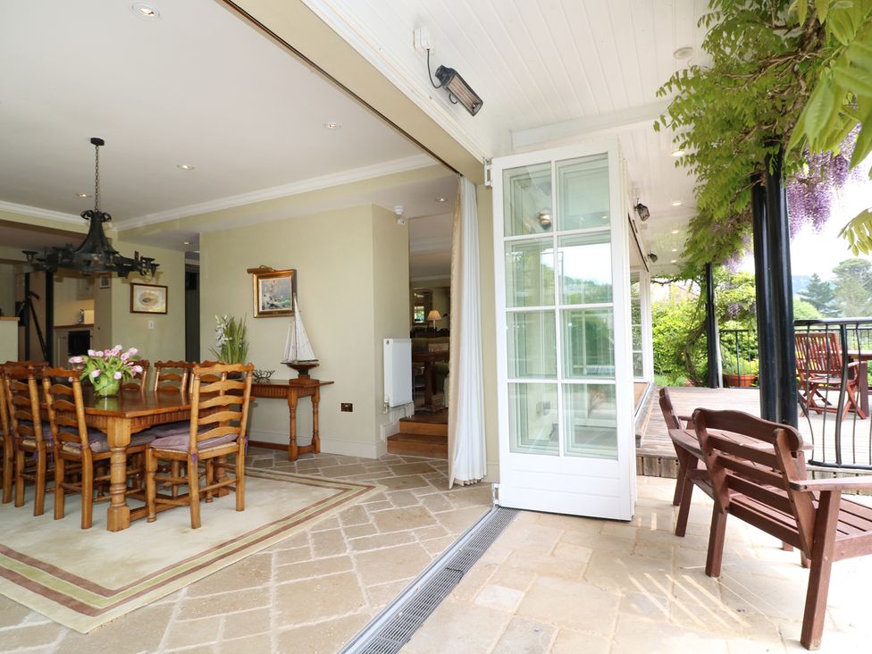A dining area with wooden table and chairs opening onto a covered patio with wooden bench and outdoor seating at Elphinstone Newton Ferrers near Plymouth
