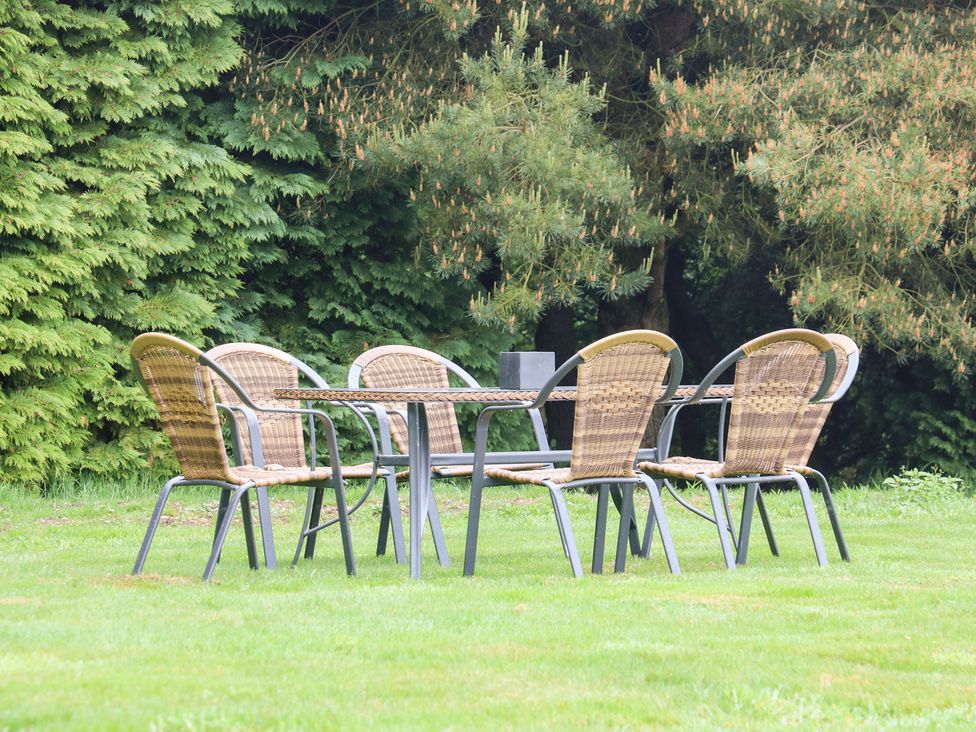 A set of six wicker chairs around a metal and wicker table on grass with large trees behind at Heron Lodge in Mavis Enderby near Spilsby