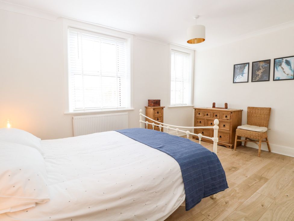 A bedroom with a white bed and blue blanket wooden dressers a wicker chair and three framed pictures at Springfield in Hempton near Fakenham