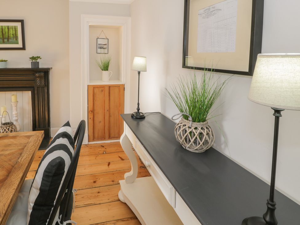 A room with a wooden floor a black and white console table with plants two table lamps a wooden dining table with black and white striped cushions a fireplace with candles and a framed picture on the wall at Bwthyn Morse in Llanfair