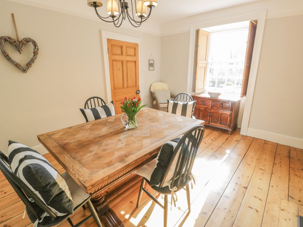 A dining room with a wooden table and four chairs with striped cushions a wooden sideboard under a window and a wicker chair next to a wooden door at Bwthyn Morse in Llanfair