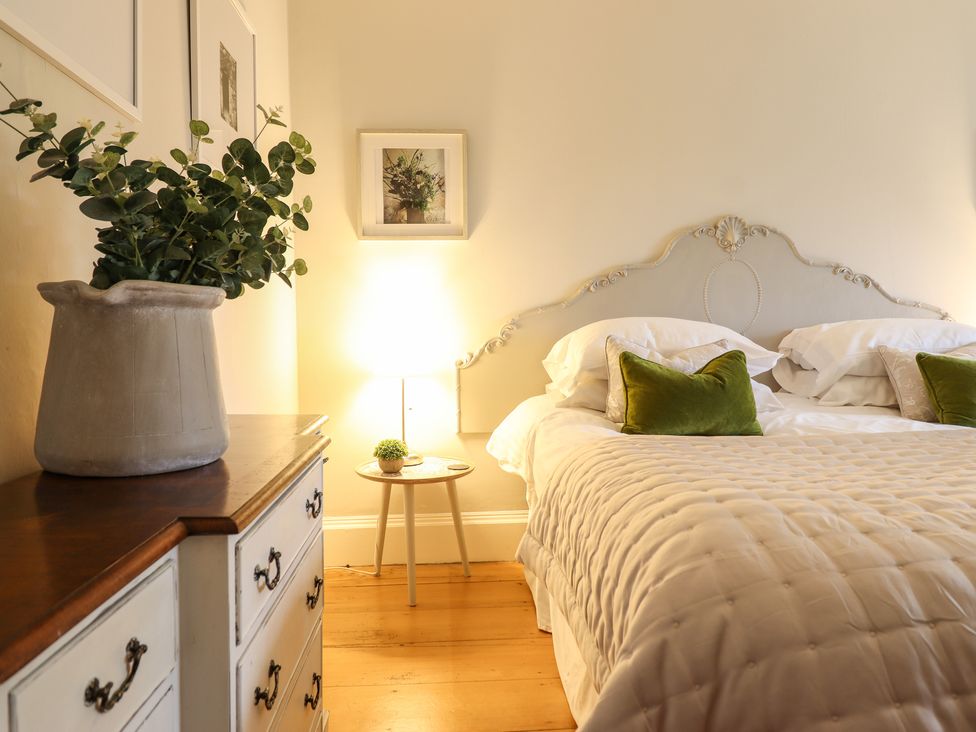 A bedroom with a bed with white bedding and green pillows a wooden dresser with a plant and a small round side table with a lamp at Bwthyn Morse Llanfair