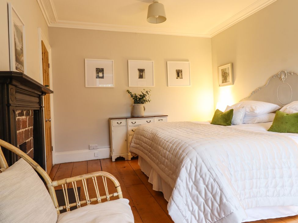 A bedroom with a bed with white bedding green pillows a wooden floor a white dresser with a vase on it framed pictures on the wall a wooden door and a fireplace at Bwthyn Morse in Llanfair