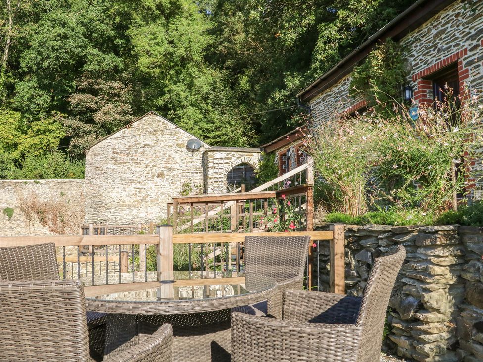An outdoor patio area with wicker chairs and a glass table in front of a stone building with plants at Rose Cottage in Barnstaple