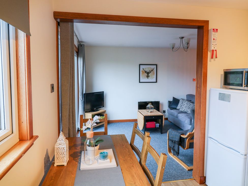 A dining area with a wooden table and chairs leading into a living room with a gray sofa a wooden coffee table a TV and wall art at Kestrel Lodge in Dumfries