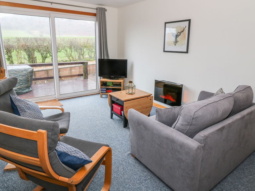 A living room with two gray chairs a gray sofa a wooden coffee table a wall-mounted fireplace a small TV on a stand and sliding glass doors showing an outside view at Kestrel Lodge in Dumfries