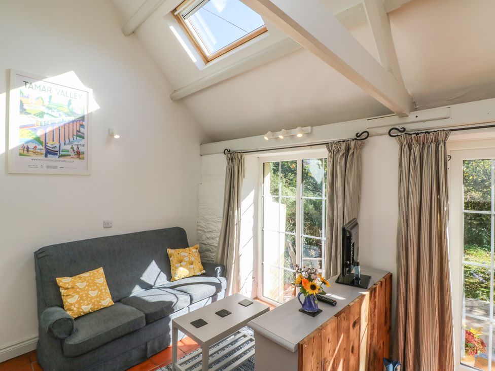 A living room with a blue sofa with yellow cushions a small white coffee table a TV on a wooden cabinet and glass doors with curtains at The Daffodil Shed in Bere Alston
