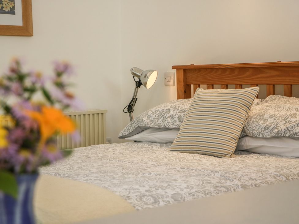 A bedroom with a wooden bed frame a striped pillow patterned bedding a metal desk lamp and flowers in a vase at The Daffodil Shed in Bere Alston