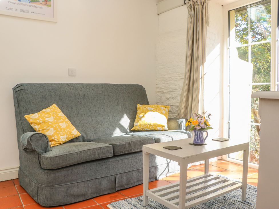 A sitting area with a gray sofa with yellow pillows a white coffee table with flowers and a glass door letting in sunlight at The Daffodil Shed in Bere Alston