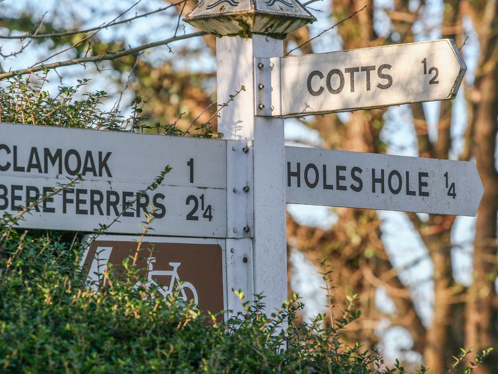 A white signpost with distances to Clamoak Bere Ferrers Cotts and Holes Hole surrounded by trees and bushes