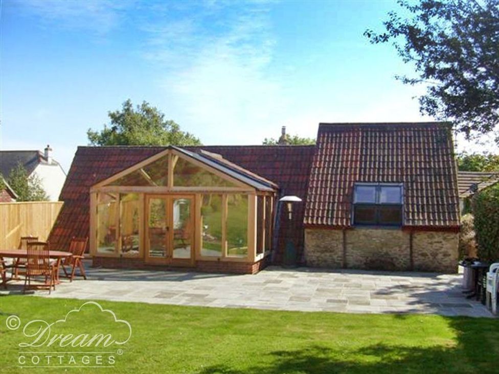 A backyard with a glass conservatory attached to a stone and tile roof building and wooden outdoor furniture on a patio at Fossil Barn in Weymouth