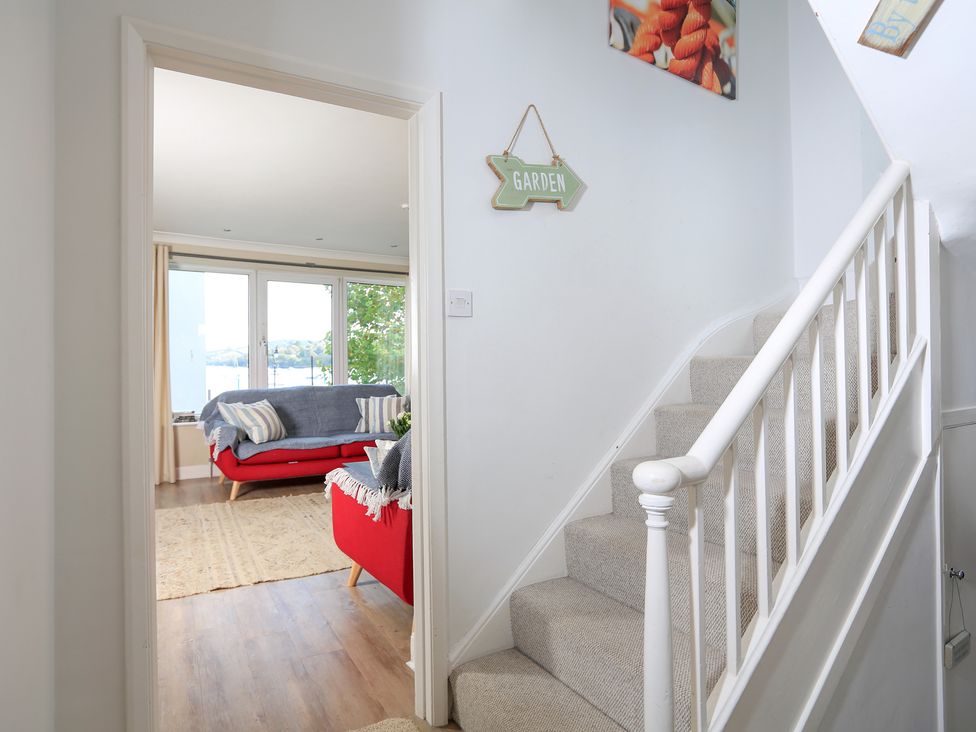 A staircase with carpet leading to a living room with red sofas and large windows overlooking water at Normandy Watch in Salcombe